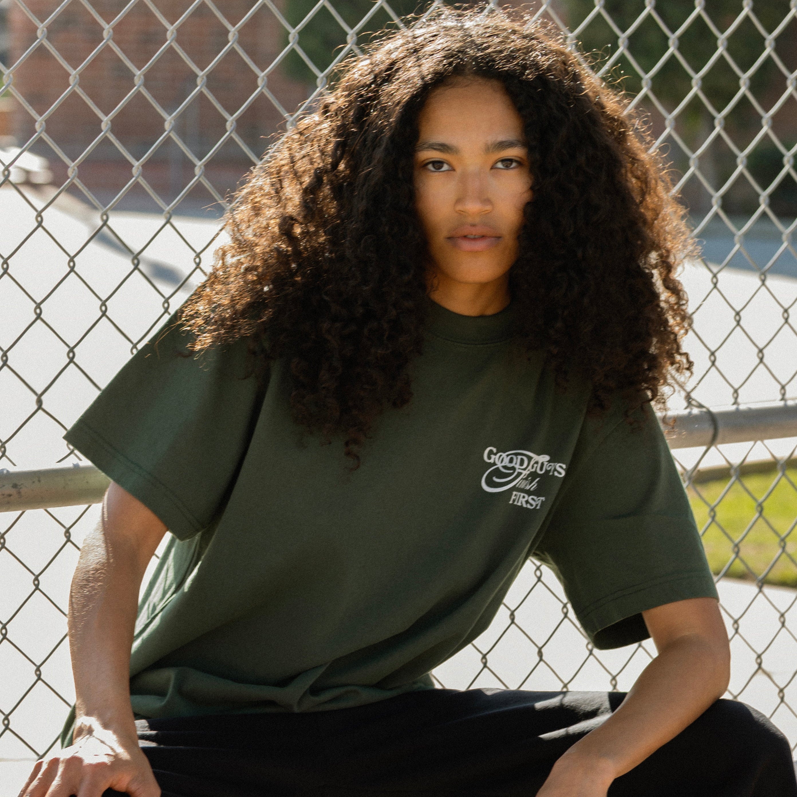 Person wearing a green t-shirt with a logo, sitting against a chain-link fence.
