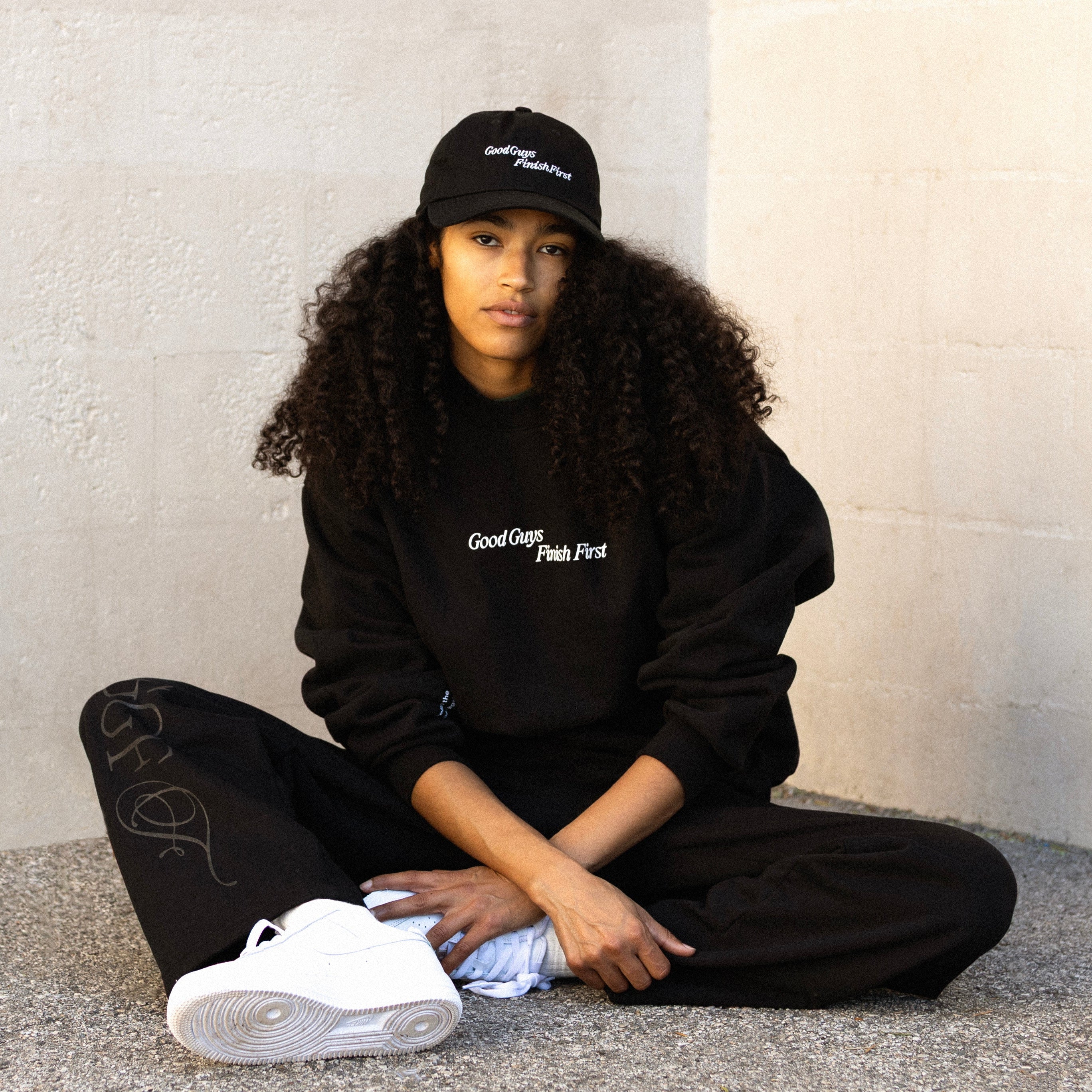 Person wearing a black outfit with text, sitting on the ground against a light-colored wall.