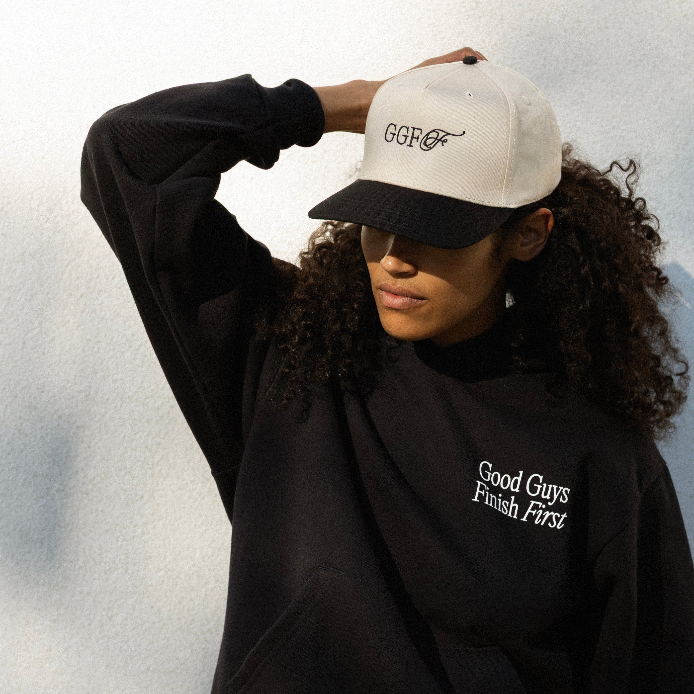 Person wearing a black sweatshirt with text and a white cap with text, against a light background